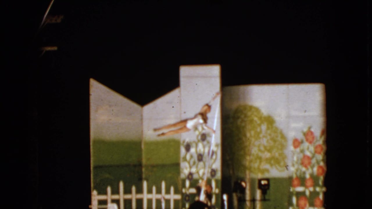 Young acrobat balanced on a pole above an assisting adult in front of a painted garden backdrop.