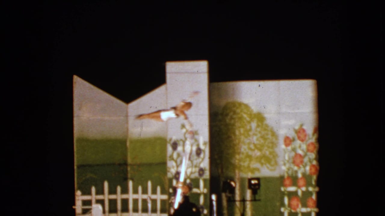 Young performer extends horizontally from a pole as an adult stands below near painted scenery.