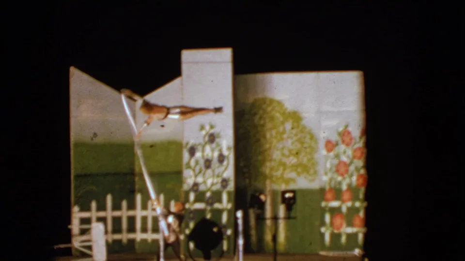 1963 Walworth, Wisconsin 8mm frame with the young acrobat and adult assistant using a long pole before painted garden scenery.