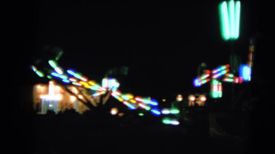 A soft, dark 8mm view of Walworth fairground lights, with colorful ride streaks and green vertical lamps.