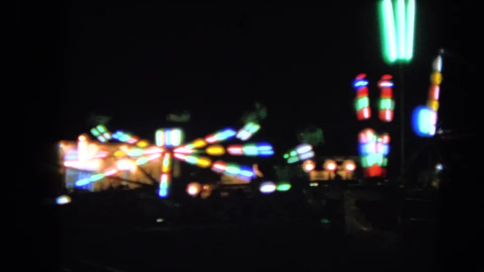 A dark 8mm frame from Walworth with bright, blurred carnival ride lighting and indistinct fairground structures.