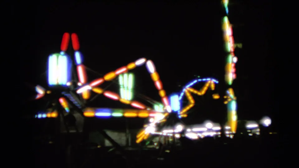 Blurry 8mm frame of a nighttime carnival ride, with colored lights and shadowed foreground forms.