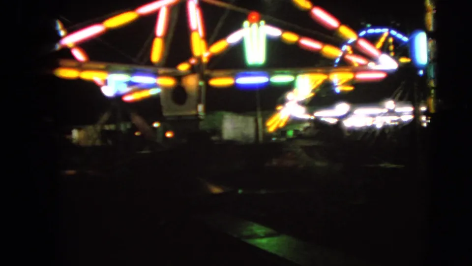 Closer 1963 Walworth carnival view showing blurred ride lighting, white bulbs, and dark fairground edges.