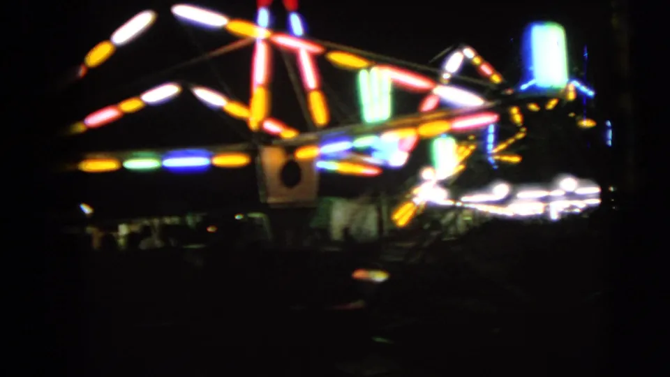 A visitor-shot 1963 fairground view shows a carnival ride's colored lights glowing and blurring against the night in Walworth, Wisconsin.