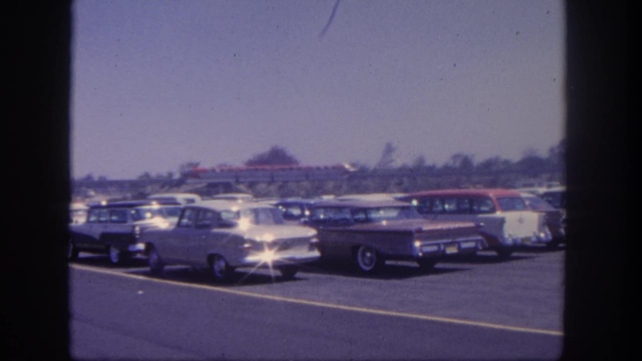 Rows of parked 1960s cars in a bright lot with distant trees and an elevated train behind them.
