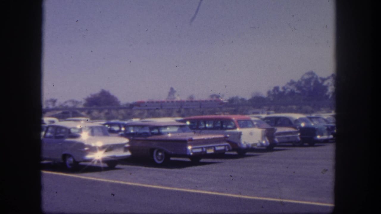 Early-1960s automobiles sit in a Disneyland parking lot, with a red monorail-like train crossing the background under a bright sky.