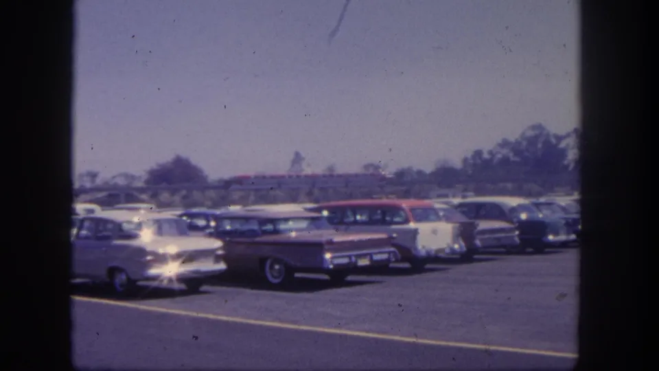 Early-1960s automobiles sit in a Disneyland parking lot, with a red monorail-like train crossing the background under a bright sky.