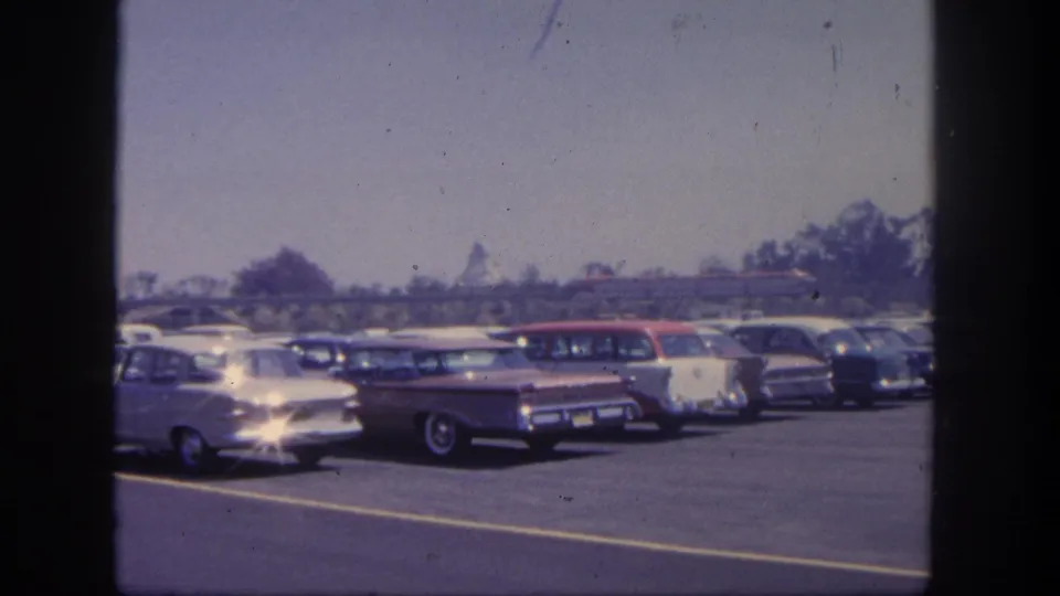 Home-movie frame of the 1962 Disneyland lot showing rows of classic American cars, a wagon near center, trees, and a red train behind the parking area.