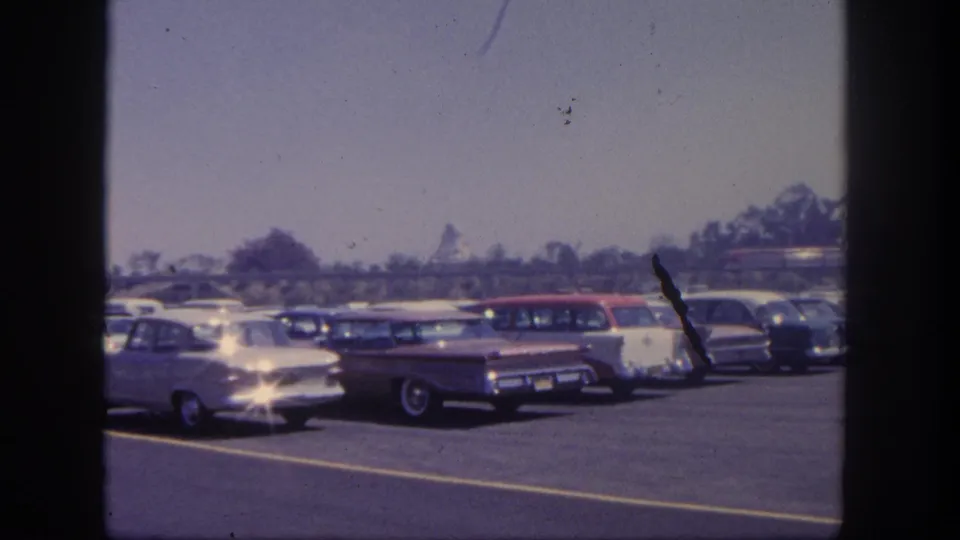 1962 Disneyland parking lot frame with classic sedans and wagons, a red train in the background, asphalt markings, and noticeable 8mm dust marks.