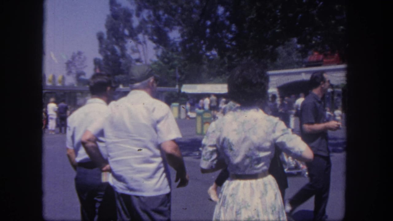 Adults walk along a shaded Disneyland pathway with buildings and trees ahead.