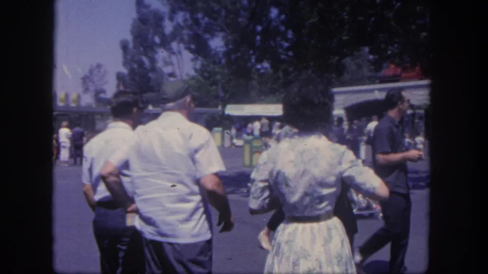 1962 Disneyland home-movie frame showing adult visitors walking from behind along a sunlit pathway toward park buildings under trees.