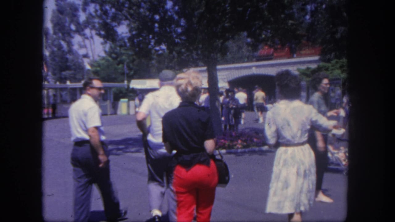 Adults stand and walk near a Disneyland entrance-area walkway under trees.
