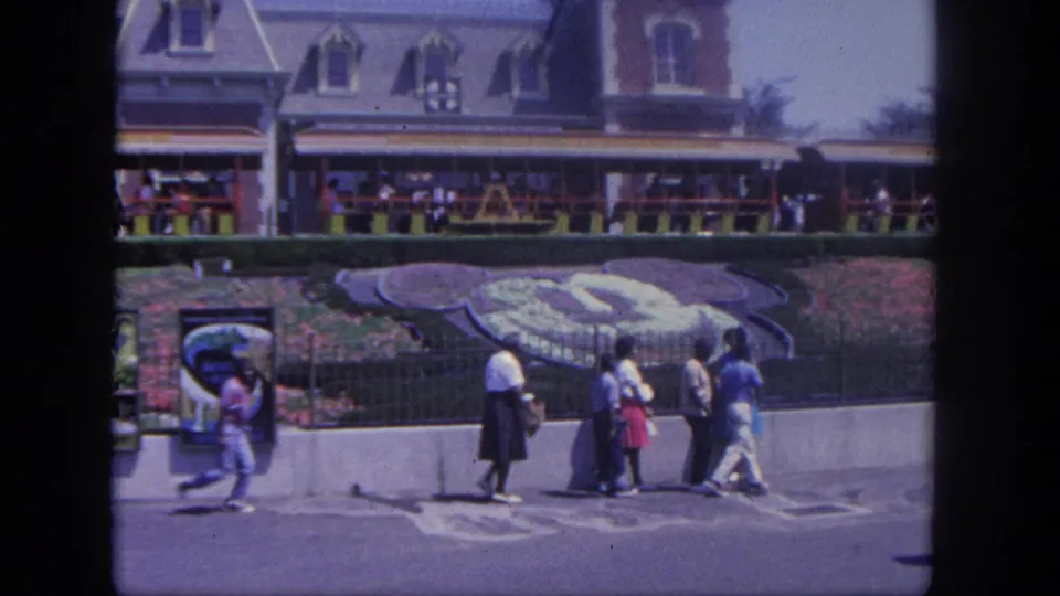 1962 Disneyland home-movie frame showing guests near a large Mickey-shaped planting bed, railings, display panels, and a building facade.