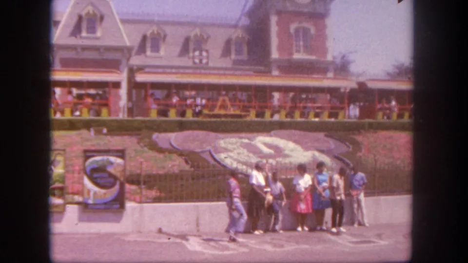 1962 Disneyland frame showing adults gathered for a photo near the Mickey-shaped floral landscaping, railing, and ornate park building facade.