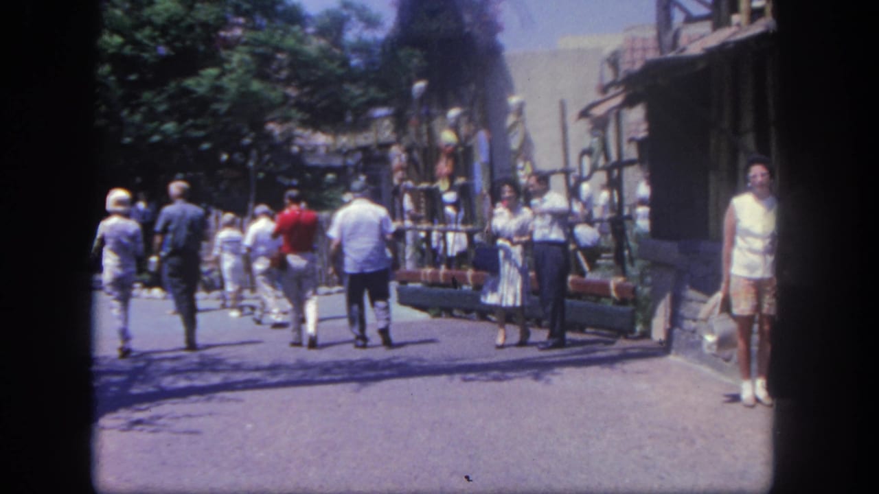 Guests walk along a sunny themed street near trees and rustic buildings.