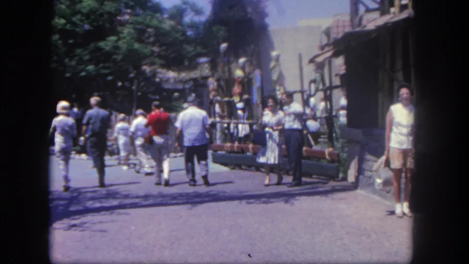 1962 Disneyland visitors walk along a themed street with trees, railings, and rustic facades visible in soft 8mm color.