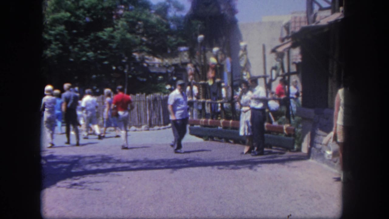 Guests in 1960s clothing walk past a fenced area and rustic Disneyland street facade, with trees casting shade nearby.