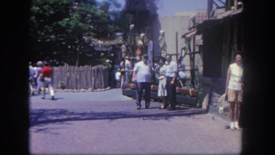 Disneyland guests walk toward the camera along a sunny 1962 themed street, with a rustic building and foliage at the side.