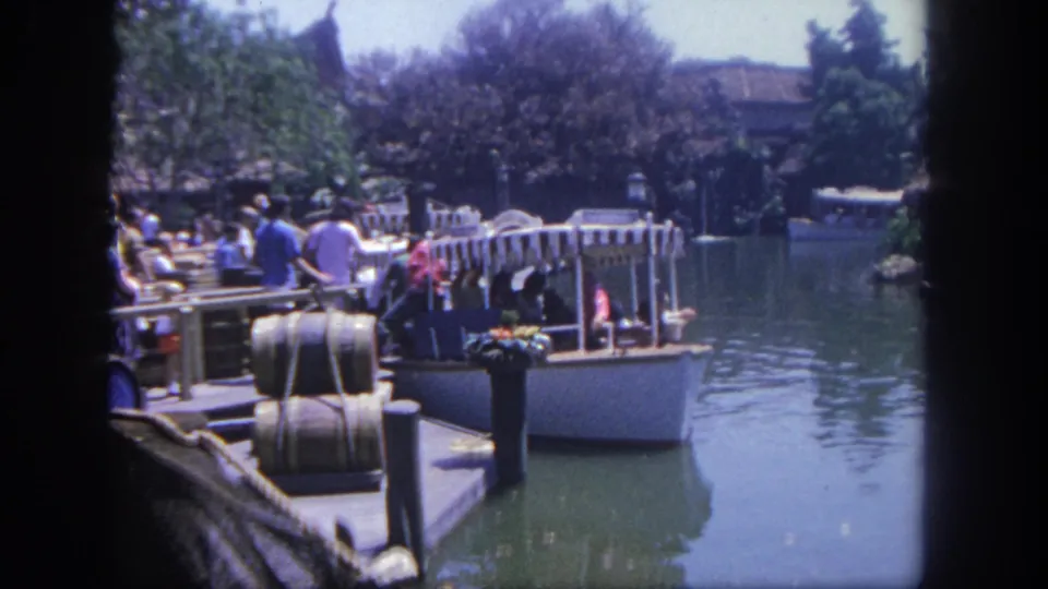 A Disneyland boat ride appears on a calm waterway with seated passengers, dock railings, trees, and visitors nearby in 8mm footage.