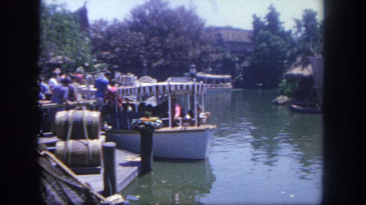 1962 Disneyland home-movie frame showing visitors on a dock beside a riverboat-like craft on a green waterway with trees and themed scenery.