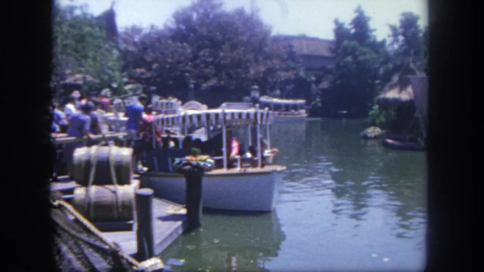 1962 Disneyland home-movie frame showing visitors on a dock beside a riverboat-like craft on a green waterway with trees and themed scenery.