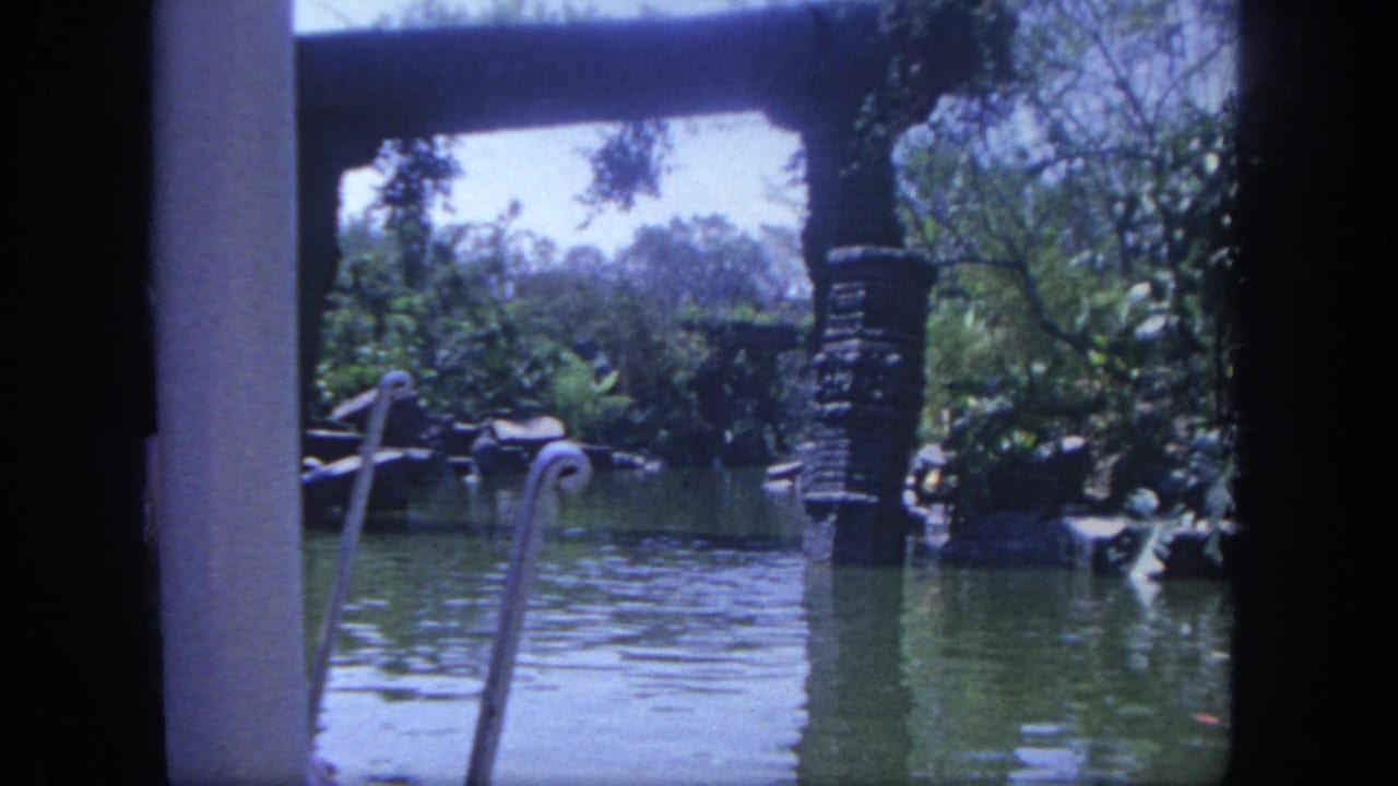 1962 Disneyland waterway view from home-movie footage, with decorative railings, a stone post, reflective water, trees, and lush planting.