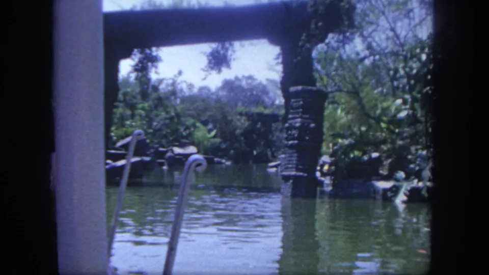 1962 Disneyland waterway view from home-movie footage, with decorative railings, a stone post, reflective water, trees, and lush planting.