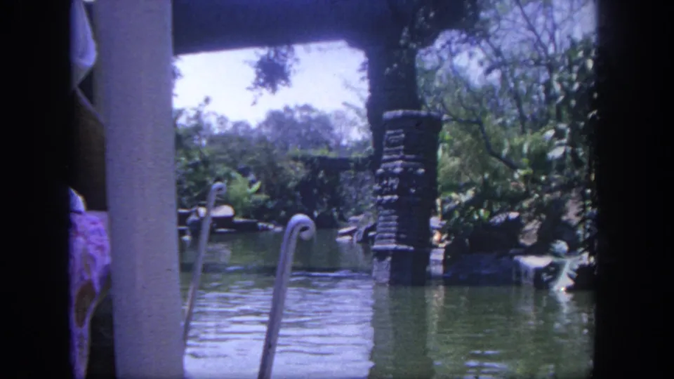 Home-movie frame at Disneyland in 1962 showing swan-neck railings, a stone post, water, trees, and tropical-style planting from a visitor viewpoint.