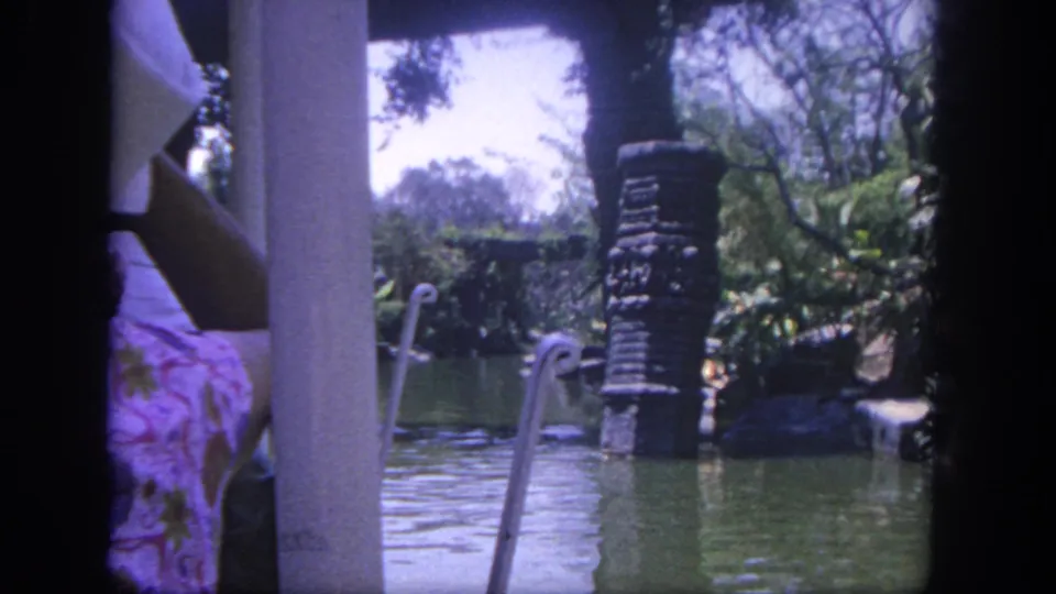1962 Disneyland home-movie frame with a partial visitor in patterned clothing near a column, looking across a green waterway with railings and foliage.