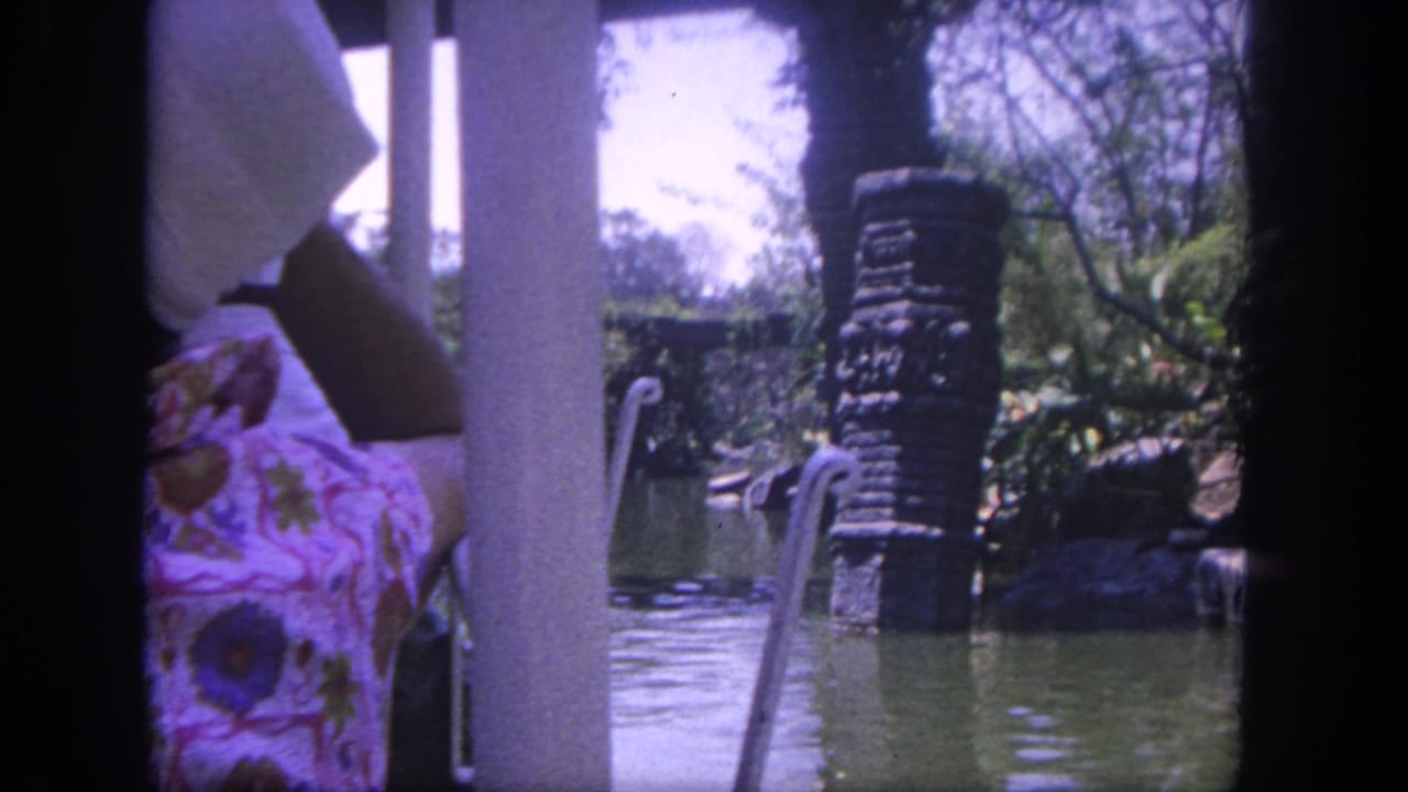Boat-side view of green water, stone pillar, foliage, and a seated passenger at the left edge.