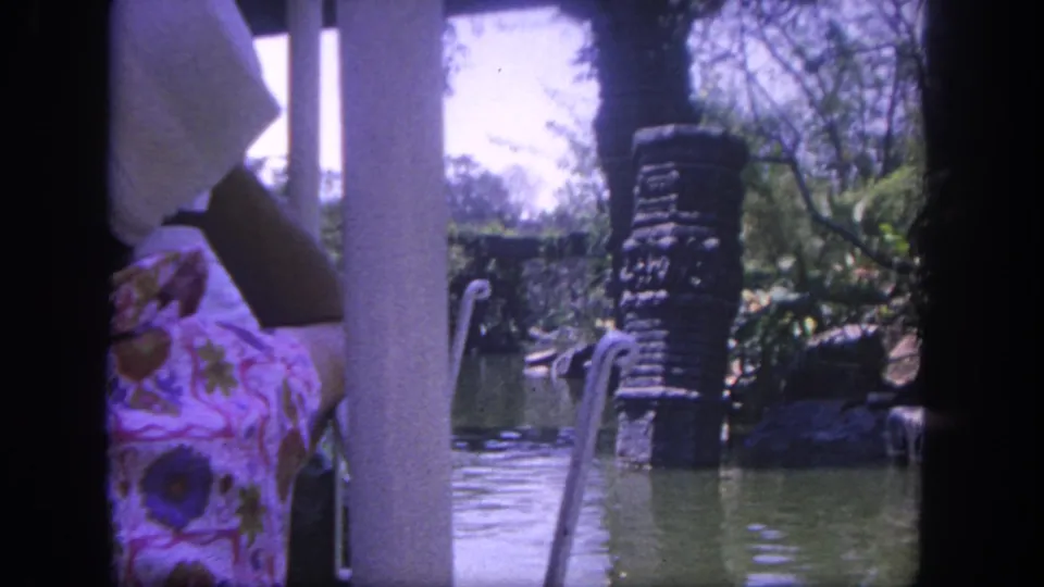 1962 Disneyland ride footage from a boat, showing green water, tropical planting, a stone pillar, railings, and a partly visible seated passenger.