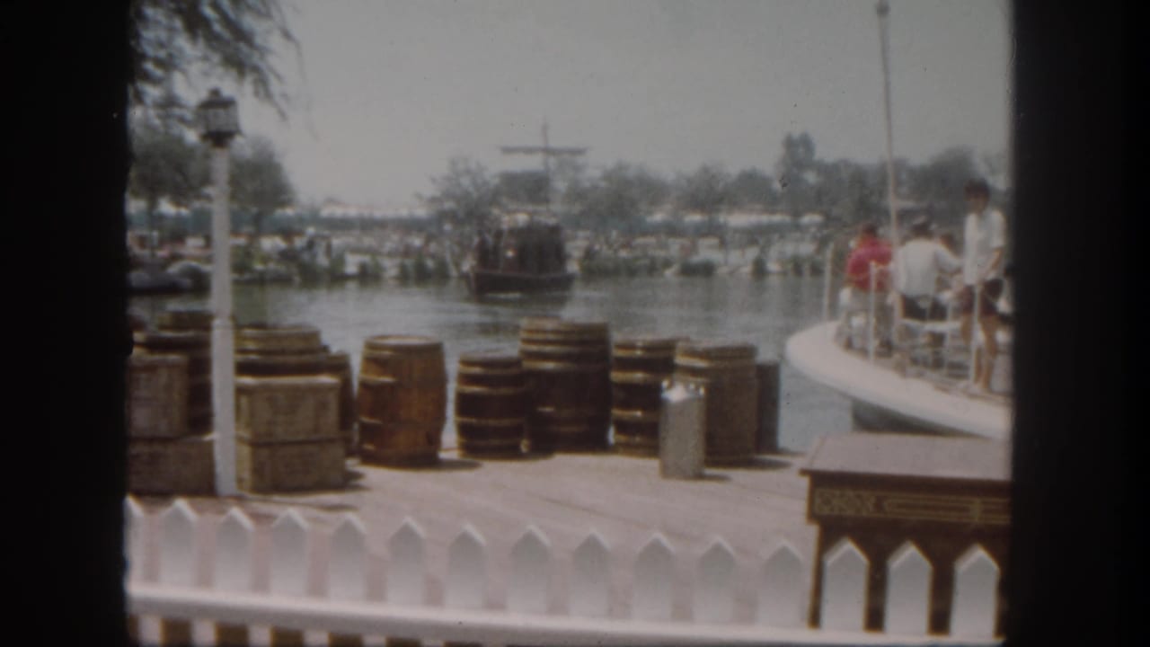 Themed dock with barrels, crates, a white railing, and a boat on the water behind it.