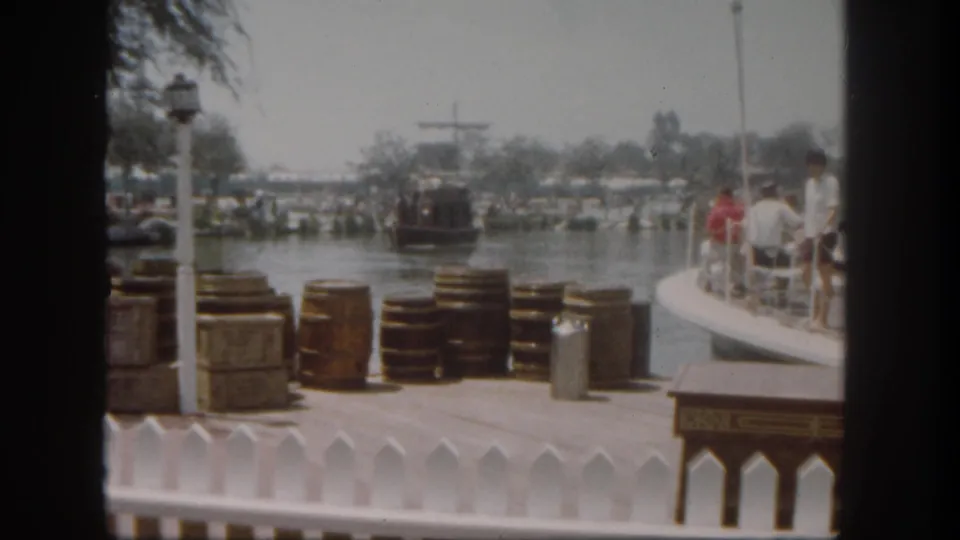 1962 Disneyland dock scene with wooden barrels and crates in front, a white railing, guests at right, and a paddlewheel-style boat on the water.