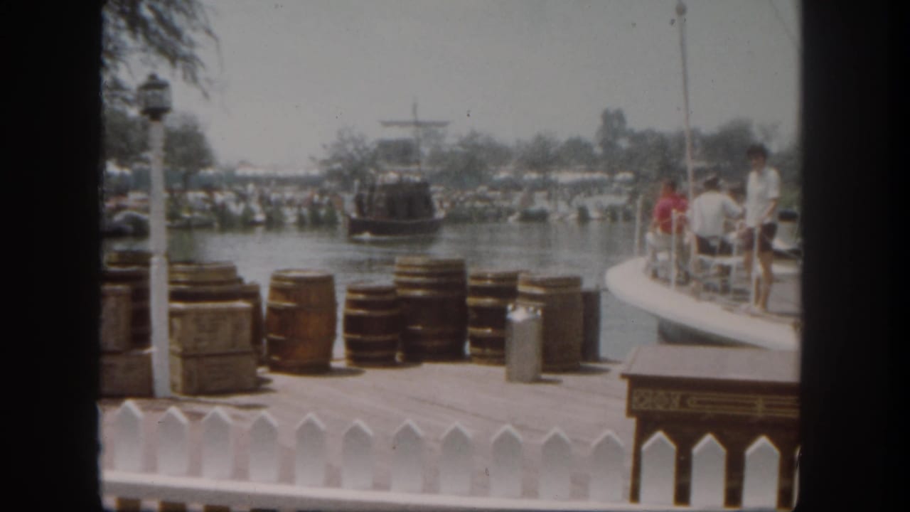 Dockside barrels and crates with guests on a curved pier watching a boat across the water.
