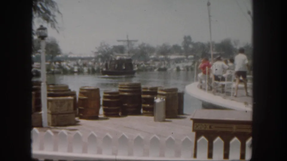 1962 Disneyland dockside home-movie frame with stacked barrels, crates, white fence trim, guests at right, and a boat moving on the water.