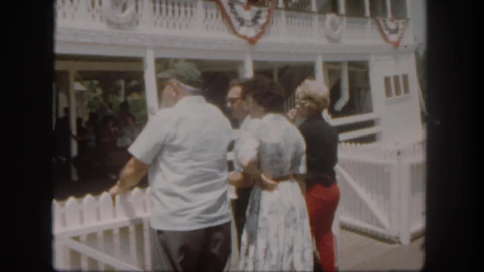 1962 Disneyland dock area with adults talking beside a white picket fence, a decorated building or boat structure, and shaded visitors behind them.