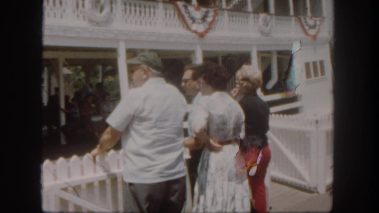 At Disneyland in 1962, adult visitors stand near a white dock fence while facing a bunting-decorated riverboat area.