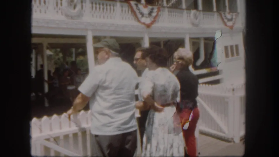 At Disneyland in 1962, adult visitors stand near a white dock fence while facing a bunting-decorated riverboat area.