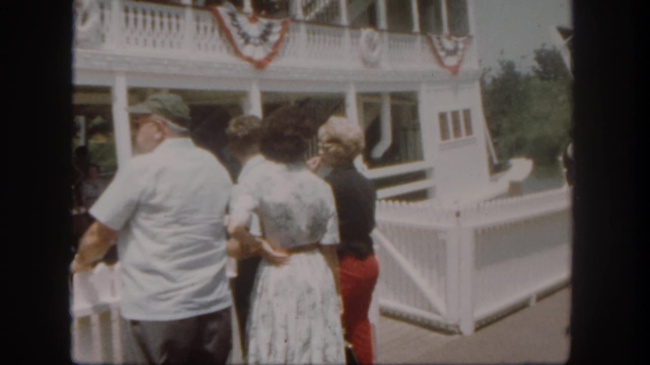 At the Disneyland riverboat dock, a man in a white shirt and two women look toward the shaded boarding area.
