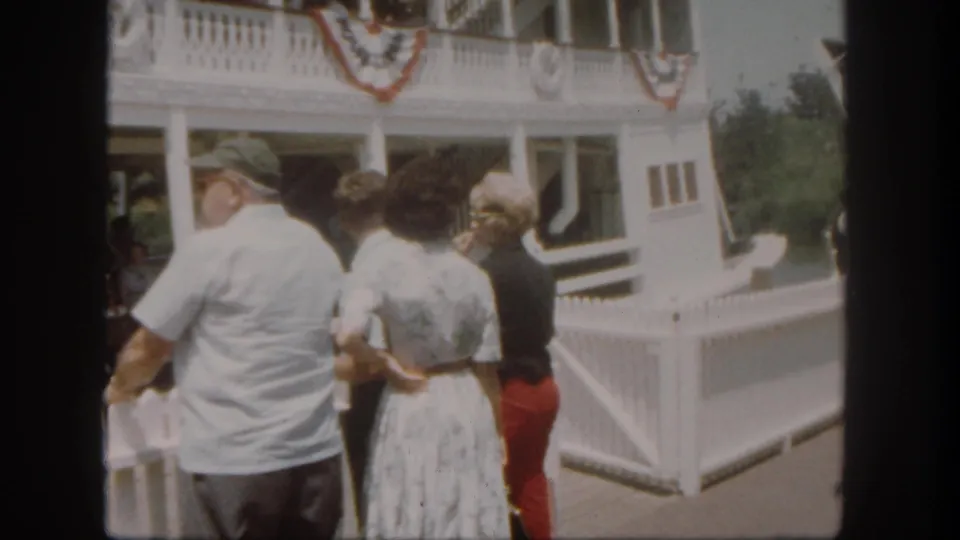 At the Disneyland riverboat dock, a man in a white shirt and two women look toward the shaded boarding area.