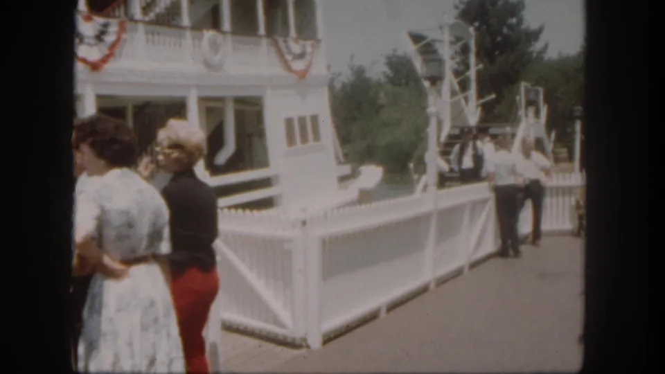 Two women remain by the Disneyland riverboat dock fence, with a gangway-like structure and trees beyond.
