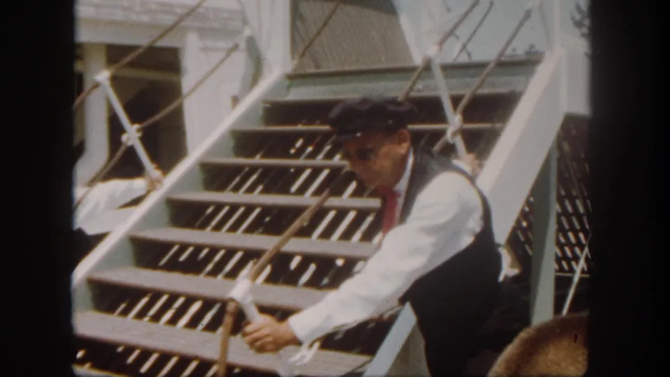 A crew member in vest and cap leans by the riverboat gangway, with white railings and stair treads filling the frame.