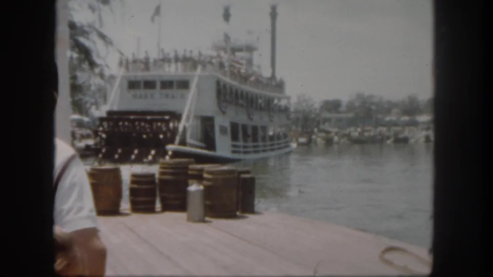 1962 Disneyland view of the Mark Twain riverboat at the dock, with guests aboard, barrels on the pier, and water beyond.