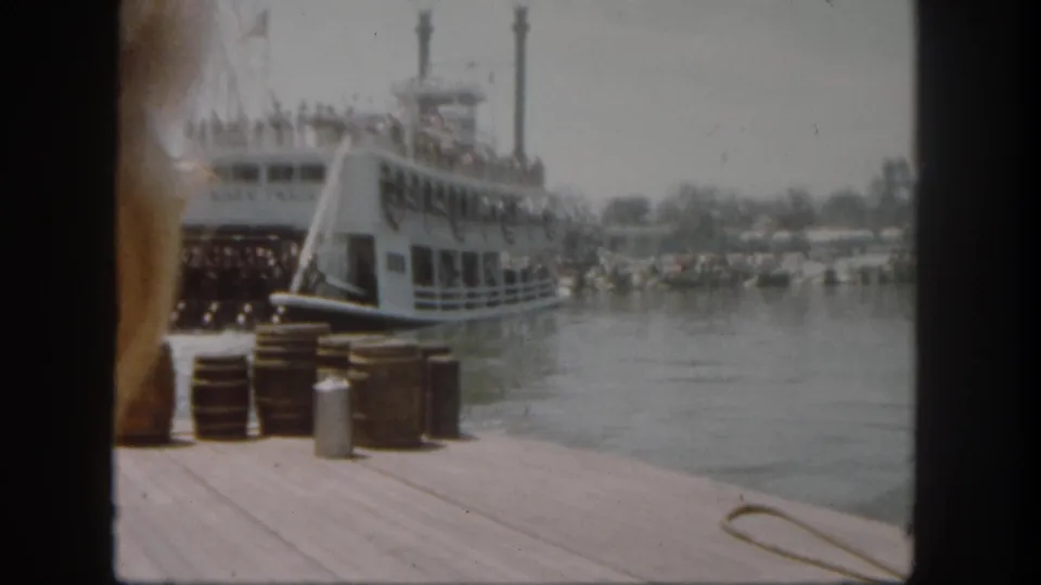 1962 Disneyland frame showing the Mark Twain riverboat by the river, dock barrels in front, and a partial foreground visitor at left.
