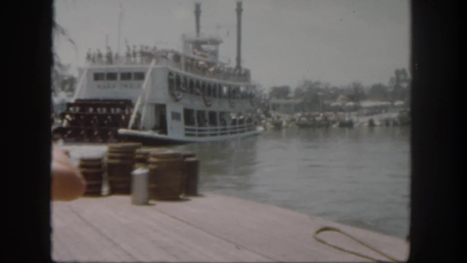 1962 Disneyland home-movie frame with the Mark Twain riverboat docked beside barrels, water, and a partial foreground arm.
