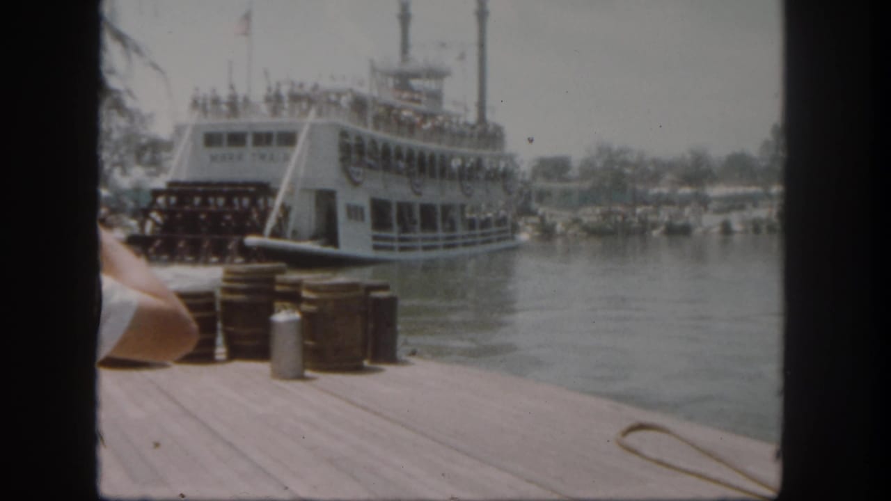 1962 Disneyland dock view of the Mark Twain Riverboat beside water, with stacked barrels, a metal can, bunting, flags, and guests aboard.