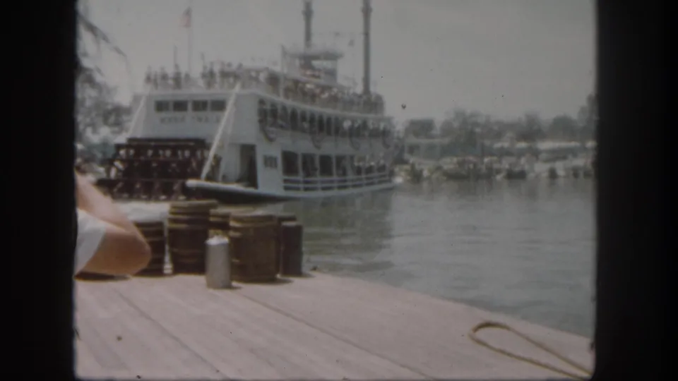 1962 Disneyland dock view of the Mark Twain Riverboat beside water, with stacked barrels, a metal can, bunting, flags, and guests aboard.