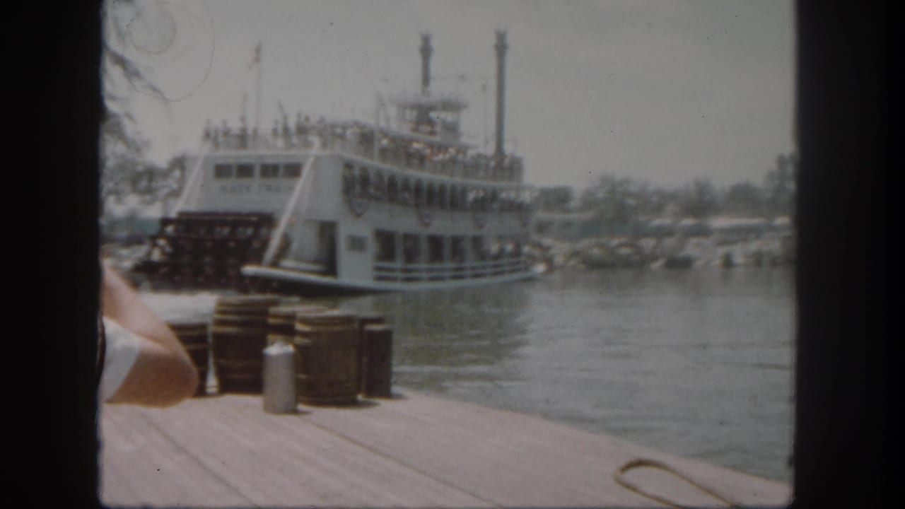 The Mark Twain Riverboat appears to move away from the Disneyland dock, with passengers on deck, flags overhead, and water between boat and camera.