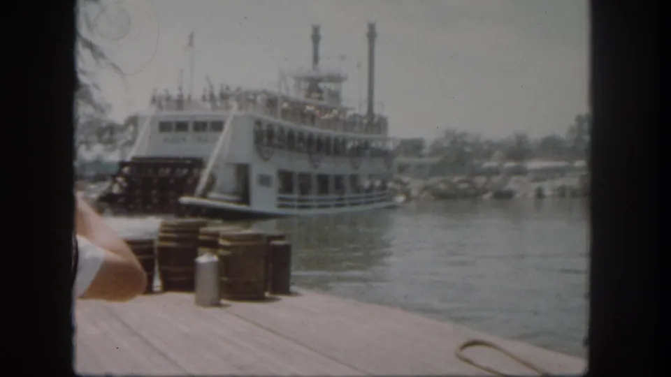 The Mark Twain Riverboat appears to move away from the Disneyland dock, with passengers on deck, flags overhead, and water between boat and camera.