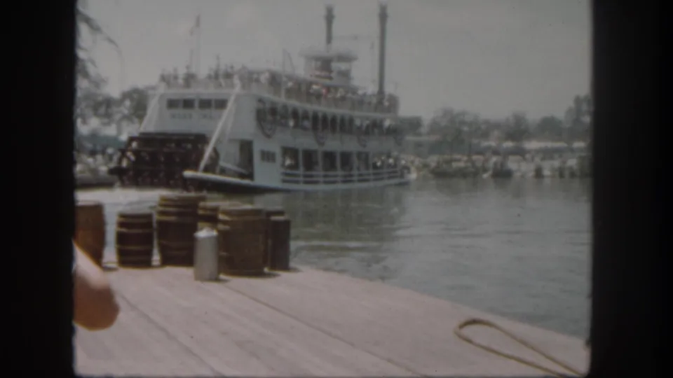 Visitor-shot 1962 Disneyland frame showing the riverboat near the dock, stacked barrels in front, guests on deck, and a partial arm entering at left.